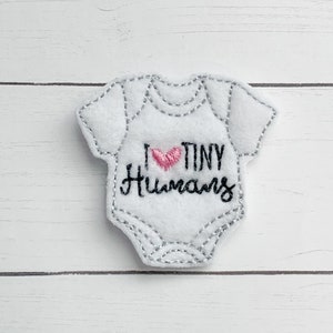 May include: White felt baby onesie applique with gray stitching. The onesie has the text "I ♥ TINY Humans" with a pink heart. The applique is on a white wooden background.