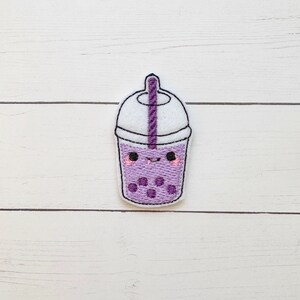 May include: A purple embroidered patch in the shape of a bubble tea drink with a smiling face. The patch features a white lid, a purple straw, and purple boba pearls. The drink has pink cheeks and black eyes.