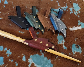 May include: A collection of handcrafted wooden wands with decorative leather sheaths in various colors, including black, green, brown, blue, and gray. Each sheath features a unique wing-like design and is adorned with metallic studs. The wands are displayed on a brown and turquoise patterned surface.