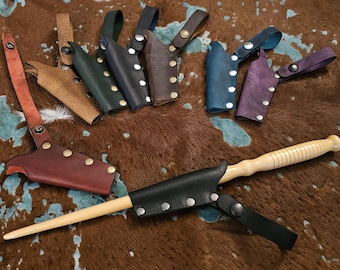 May include: A set of nine leather sheaths for wands or other small objects. The sheaths are in various colors, including brown, black, blue, and purple. The sheaths are decorated with silver rivets. A wooden wand is also pictured.