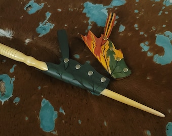 May include: Two leather sheaths for a wand or staff. One sheath is green with a leafy design and the other is multi-colored with a leaf design. Both sheaths have metal snaps.