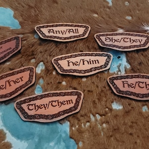 Puede incluir: Siete etiquetas de nombre de cuero con diferentes opciones de pronombres, que incluyen "Pide mis pronombres", "Cualquiera/Todos", "Él/Él", "Ella/Ella", "Ellos/Ellos", "Ella/Ellos" y "Él/Ellos".