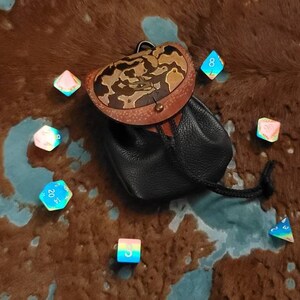 May include: A black leather dice bag with a brown and tan leather top, surrounded by iridescent dice. The bag has a drawstring closure and a decorative leather design. The dice are various colors.