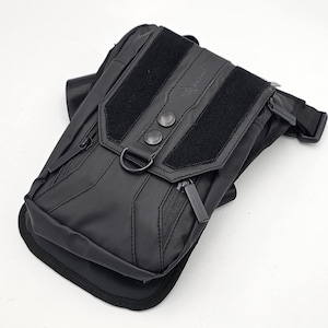 May include: Black tactical leg bag with multiple zippered compartments and adjustable straps. Features two Velcro patches and two decorative buttons on the flap. Includes a metal D-ring and a black strap for secure wear.