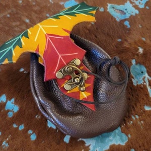 Op de afbeelding: Een bruin leren tasje met een decoratieve blad-vormige flap in herfstkleuren. De flap heeft rode, gele en groene bladeren. Een bronzen sluiting sluit het tasje af, en een zwart trekkoord is bevestigd.