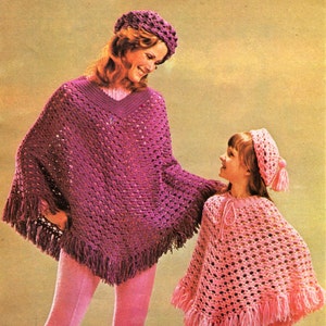 May include: Two women wearing crocheted ponchos and hats. The woman on the left is wearing a purple poncho with a purple hat, and the woman on the right is wearing a pink poncho with a pink hat. Both ponchos have fringe.