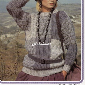 May include: A grey and white chunky knit jumper with a cable knit pattern. The jumper has a round neckline and long sleeves. The model is wearing a grey hat and a silver chain necklace. The jumper is 76-112 cm (30-44 inches) in size.