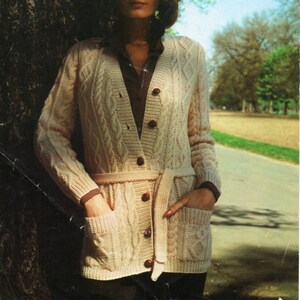 May include: A woman wearing a cream-colored cable knit cardigan with a brown belt and buttons. The cardigan has pockets and is worn over a brown shirt. The text "PATONS Totem Double Crepe 34-40 in. 2445 8p" is visible in the top right corner of the image.