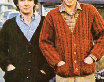 May include: Two men pose in front of a vintage car. One wears a dark brown cardigan over a light blue collared shirt. The other wears a red-orange cardigan over a plaid shirt. Both cardigans have pockets.