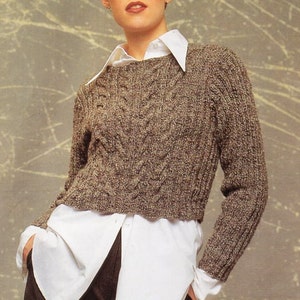 Könnte beinhalten: Ein brauner Zopfmusterpullover mit Rundhalsausschnitt. Der Pullover ist kurz und hat lange Ärmel. Das Model trägt den Pullover zu einem weißen Hemd mit Knöpfen und einer schwarzen Hose. Der Text "Knit this from just 5x50 gram balls!" ist im Bild zu sehen.