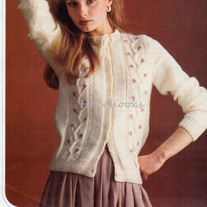 K&ouml;nnte beinhalten: Ein wei&szlig;er Strickcardigan mit einem Zopfmuster und rosa Blumen Stickerei. Der Cardigan hat einen Knopfverschluss und wird an einem Model mit einem braunen Rock gezeigt. Die Musternummer ist 588.