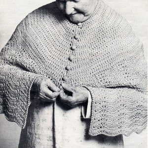 May include: A close-up of a woman wearing a crocheted shawl with buttons. The shawl is a light gray color and has a scalloped edge. The woman is wearing a white dress underneath the shawl.
