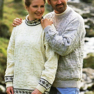 Pode incluir: Um casal usando suéteres de malha. O suéter da mulher é creme com um padrão de floco de neve e detalhes em preto. O suéter do homem é cinza com um design texturizado. Ambos usam jeans azuis.