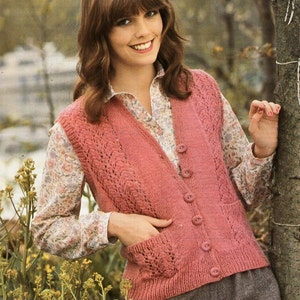 May include: A pink knitted waistcoat with a lace pattern and button fastening. The waistcoat has pockets on the sides. The waistcoat is modelled by a woman with brown hair and a floral blouse.