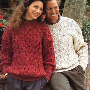Könnte beinhalten: Ein Paar trägt passende Strickpullover mit einem Zopfmuster. Die Frau trägt einen roten Pullover und der Mann einen weißen Pullover. Sie stehen vor einer Backsteinmauer.