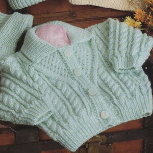 May include: A set of knitted baby clothes in cream and light green. The set includes a hooded sweater, a cardigan, a hat, and mittens. The sweater has a cable knit pattern and the cardigan has a button closure. The hat and mittens are also cable knit.