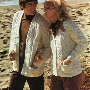 May include: Two people wearing white knitted cardigans with cable knit patterns. The cardigan has a button closure and a pocket on each side. The text "Patons Aran Style Capstan 34-44 in." is visible in the image.