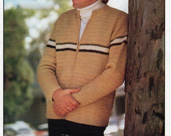 May include: A tan crocheted zip-up sweater with white and green stripes on the sleeves. The sweater is being worn by a person with blonde hair standing next to a tree. The text "Marriner Bouquet DK 8p" is printed on the top of the image.