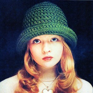 May include: A woman with long, wavy, blonde hair is wearing a green crocheted hat and a white turtleneck sweater with a silver heart-shaped pendant necklace.