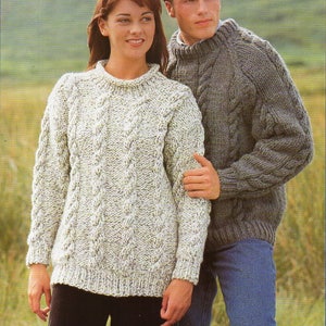 May include: A couple wearing matching cable knit sweaters. The woman's sweater is white with a cream color and the man's sweater is gray. They are standing in a field with green grass.