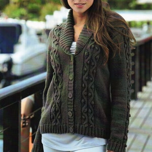 Puede incluir: Una mujer lleva un cárdigan de punto verde con cable y un gorro de lentejuelas plateadas. El cárdigan tiene un cuello ancho y botones en la parte delantera. La mujer está de pie en una pasarela de madera.