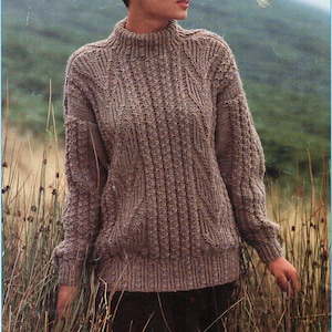 May include: A brown knitted jumper with a high neck and cable knit pattern. The jumper is worn by a person standing in a field of tall grass.
