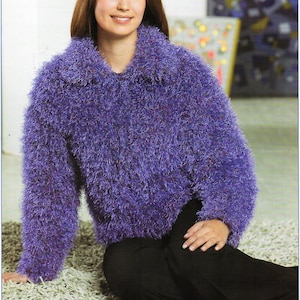 Puede incluir: Una mujer con un suéter morado y difuso con cuello y mangas largas. El suéter tiene una apariencia texturizada y se combina con pantalones negros. La mujer está sentada en una alfombra de color claro.