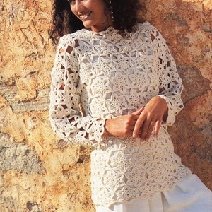 Pode incluir: Um top branco de crochê com um padrão em forma de estrela. O top tem mangas compridas e um decote redondo. A modelo está usando o top com calças brancas.