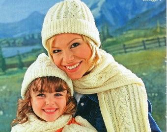 May include: A woman and a child wearing matching white knitted hats, scarves, and mittens. The hats and scarves feature a cable knit pattern. The design is labeled "Design 1".