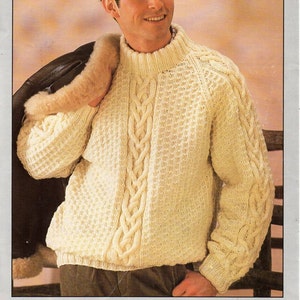 Puede incluir: Un suéter de punto blanco con un patrón de punto trenzado. El suéter tiene cuello alto y mangas largas. El suéter lo lleva puesto una persona con una chaqueta de cuero.