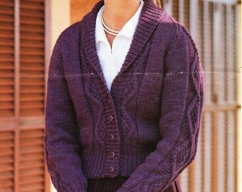 May include: A woman models a chunky, deep purple knit cardigan. The cardigan has a shawl collar, cable knit details, and button fastenings. She is also wearing a white collared shirt and a pearl necklace.