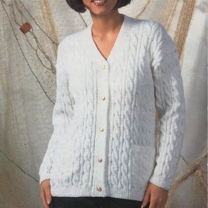 May include: White cable knit cardigan with a V-neck and button closure. The cardigan has a pocket on the left side and is shown on a model against a background of fishing net.
