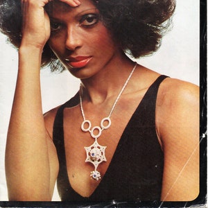 May include: A woman with dark curly hair is wearing a black sleeveless top and a white crocheted necklace with a star-shaped pendant. The pendant has blue gemstones.