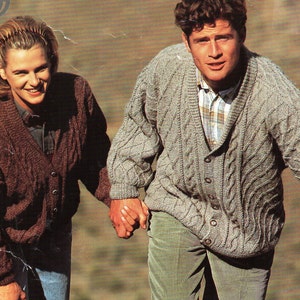 Pode incluir: Um casal vestindo cardigãs de tricô. O homem está vestindo um cardigã cinza claro com um padrão de tricô de cabo e uma camisa de colarinho por baixo. A mulher está vestindo um cardigã marrom com um padrão de tricô de cabo semelhante. Eles estão caminhando por um caminho rochoso.