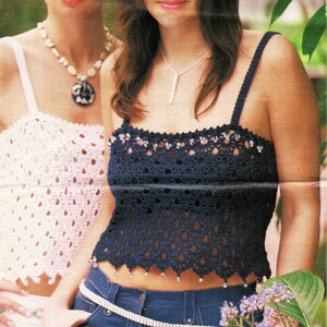 May include: Two women wearing crocheted tops. The woman on the left is wearing a white crocheted top with a blue and white pendant necklace. The woman on the right is wearing a dark blue crocheted top with a silver necklace and a silver belt.