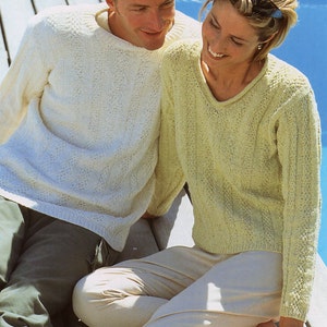 May include: A couple sitting on a wooden jetty wearing knitted jumpers. The man is wearing a white jumper and the woman is wearing a light green jumper.