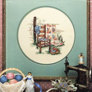 May include: A framed cross-stitch design of a chair and quilt with a doll and a sewing machine in the foreground. The cross-stitch design is in a circle with a light blue background. The doll is wearing a blue dress and has brown hair. The sewing machine is black and has a spool of thread on it.