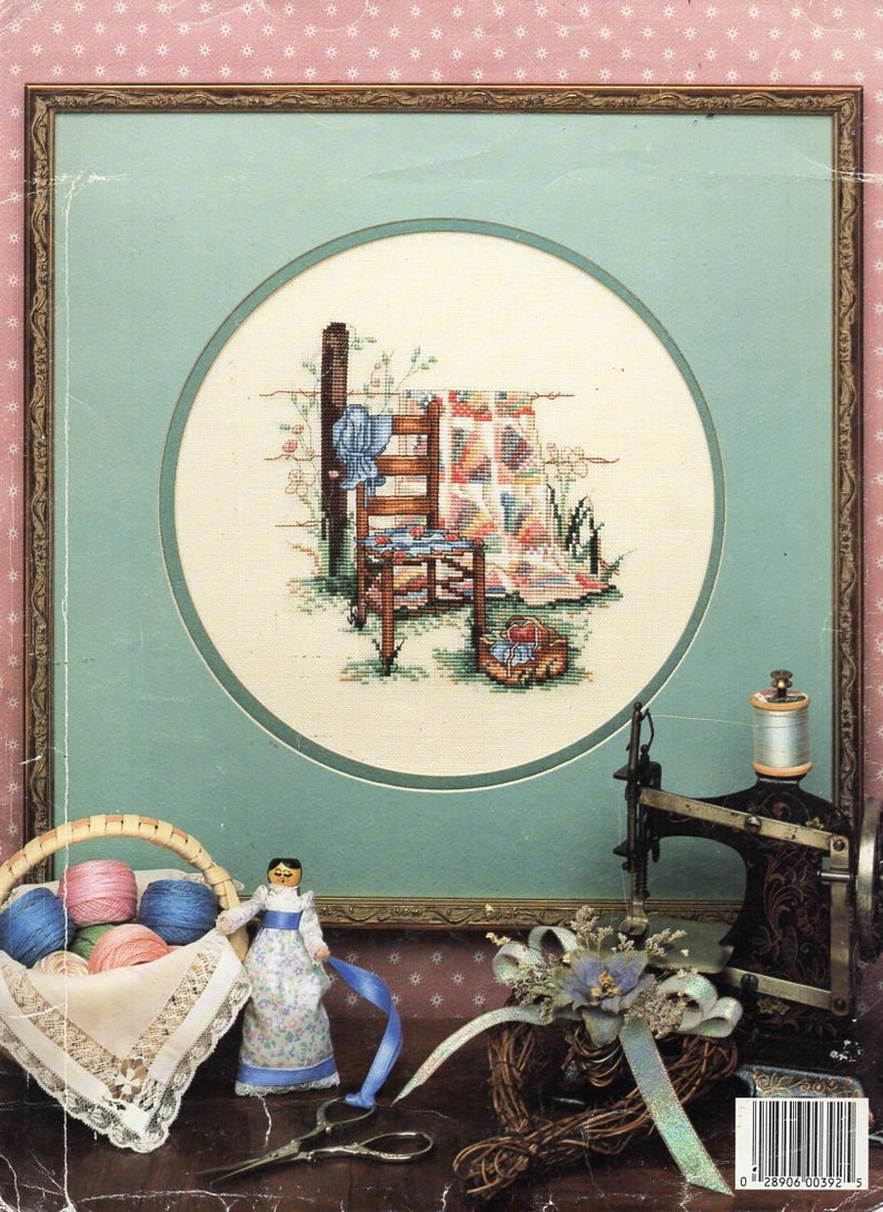 May include: A framed cross-stitch design of a chair and quilt with a doll and a sewing machine in the foreground. The cross-stitch design is in a circle with a light blue background. The doll is wearing a blue dress and has brown hair. The sewing machine is black and has a spool of thread on it.