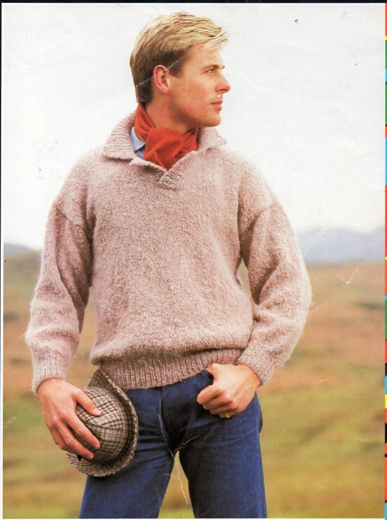 K&ouml;nnte beinhalten: Ein Mann tr&auml;gt einen hellbraunen Pullover mit Knopfleiste, einen roten Schal und eine blaue Jeans. Er h&auml;lt einen braunen Hut in seiner linken Hand.