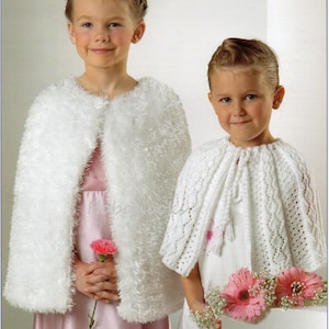 May include: Two girls wearing white knitted capes. The girl on the left is wearing a fluffy white cape and a pink dress. The girl on the right is wearing a lacy white cape and a white dress with pink flowers. Both girls are holding flowers.