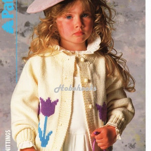 May include: A young girl wearing a light yellow cardigan with purple and blue flower designs. The cardigan has a button closure and is knitted. The girl is wearing a white shirt underneath and a pink beret. The cardigan is labeled "Easy Knit" and has a price code of "C 3315 56-71 cm 22-28 in".