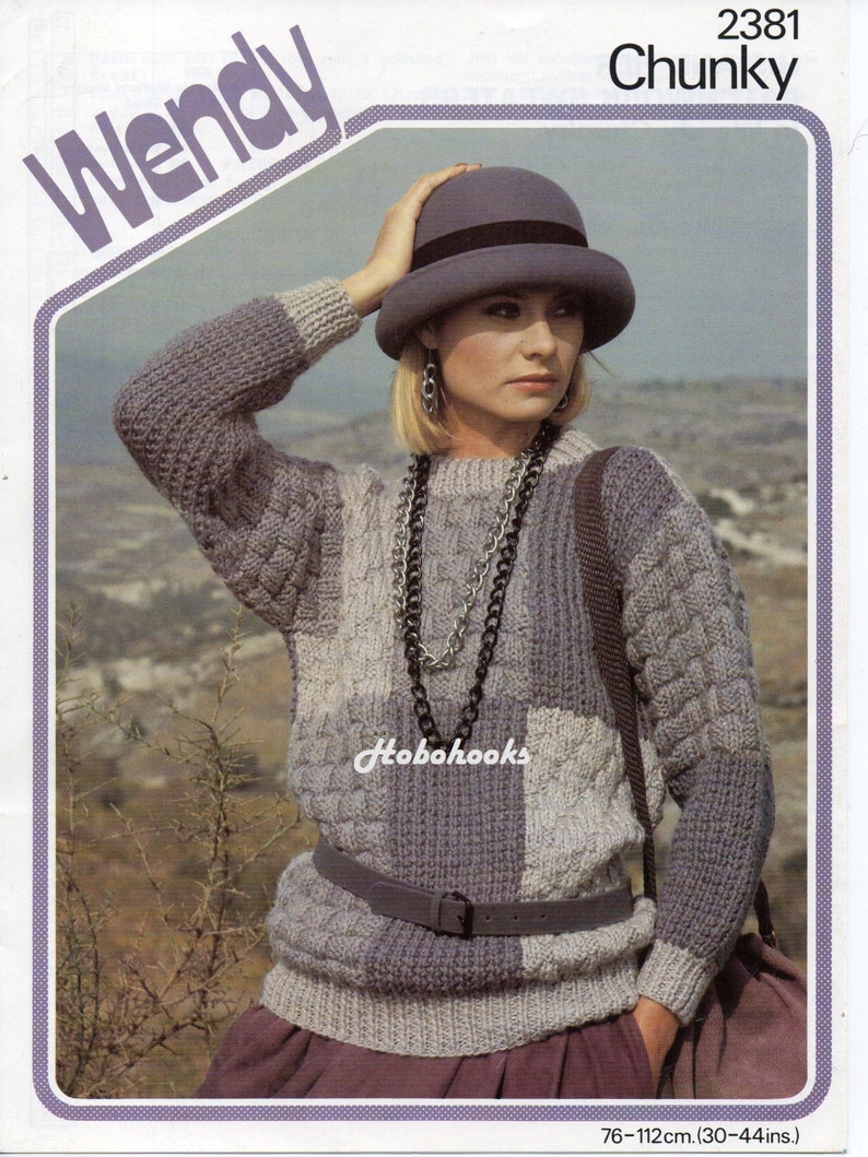 Op de afbeelding: Een grijze en witte grove gebreide trui met een kabelbreipatroon. De trui heeft een ronde hals en lange mouwen. Het model draagt een grijze hoed en een zilveren kettingketting. De trui is 76-112 cm (30-44 inch) in maat.