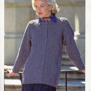 May include: A long-sleeved, gray cardigan with a cable knit pattern and button closure. The cardigan is worn over a blue shirt with a patterned scarf.