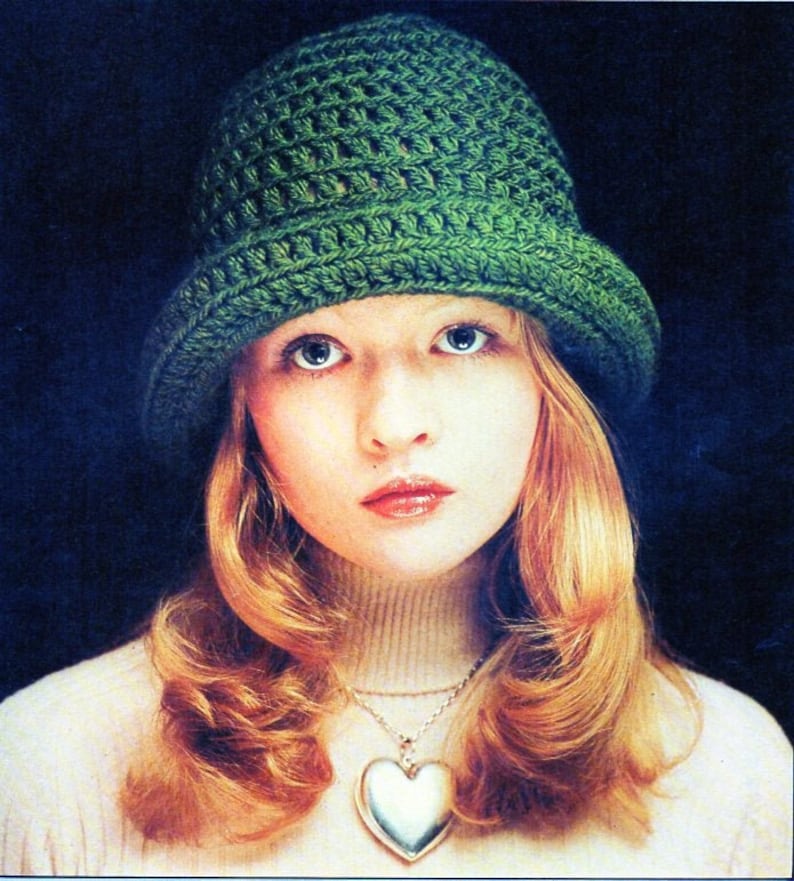 May include: A woman with long, wavy, blonde hair is wearing a green crocheted hat and a white turtleneck sweater with a silver heart-shaped pendant necklace.
