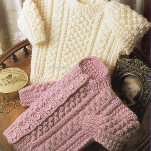 Könnte beinhalten: Zwei gestrickte Baby-Pullover, einer weiß und einer rosa, mit einem Zopfmuster. Der rosa Pullover hat Knöpfe vorne.