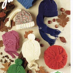 Könnte beinhalten: Eine Sammlung von sechs gestrickten Mützen in verschiedenen Farben und Stilen, darunter eine blaue Mütze mit passendem Schal, eine weiße Mütze mit Bändern, eine rosa Mütze mit Bändern, eine grüne Mütze mit Bommel, eine rote Mütze und eine mehrfarbige Mütze mit einem langen weißen Zopf. Die Mützen sind auf einem Hintergrund aus braunem Holz und Herbstlaub angeordnet. E953 Aran 8 Monate - 10 Jahre.