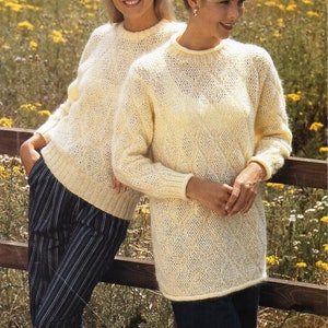 Könnte beinhalten: Zwei Frauen tragen cremefarbene Strickpullover mit einem Zopfmuster. Die Pullover sind langärmelig und haben einen runden Ausschnitt. Die Frauen stehen vor einem Holzzaun mit einem Feld voller gelber Blumen dahinter. Der Text "double knit" ist in der oberen linken Ecke des Bildes sichtbar.