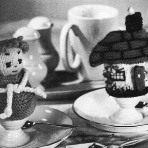 May include: Two knitted egg cozies, one shaped like a house and the other like a person, are displayed on white saucers. The house cozy has a brown roof and white walls with a window and door. The person cozy is wearing a brown coat and has a white face.