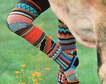 Peut inclure: Une paire de jambières en crochet colorées avec une variété de motifs et de couleurs, notamment noir, rouge, bleu, vert et blanc. Les jambières sont portées sur les jambes d'une vache debout dans un champ herbeux.
