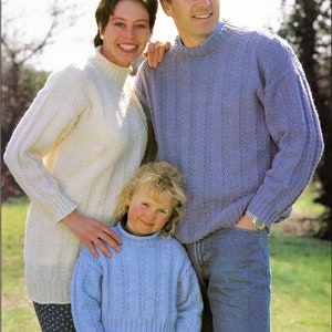 May include: A family of three wearing knitted jumpers in a variety of colours. The jumpers have a textured pattern. The family are standing in a grassy field.